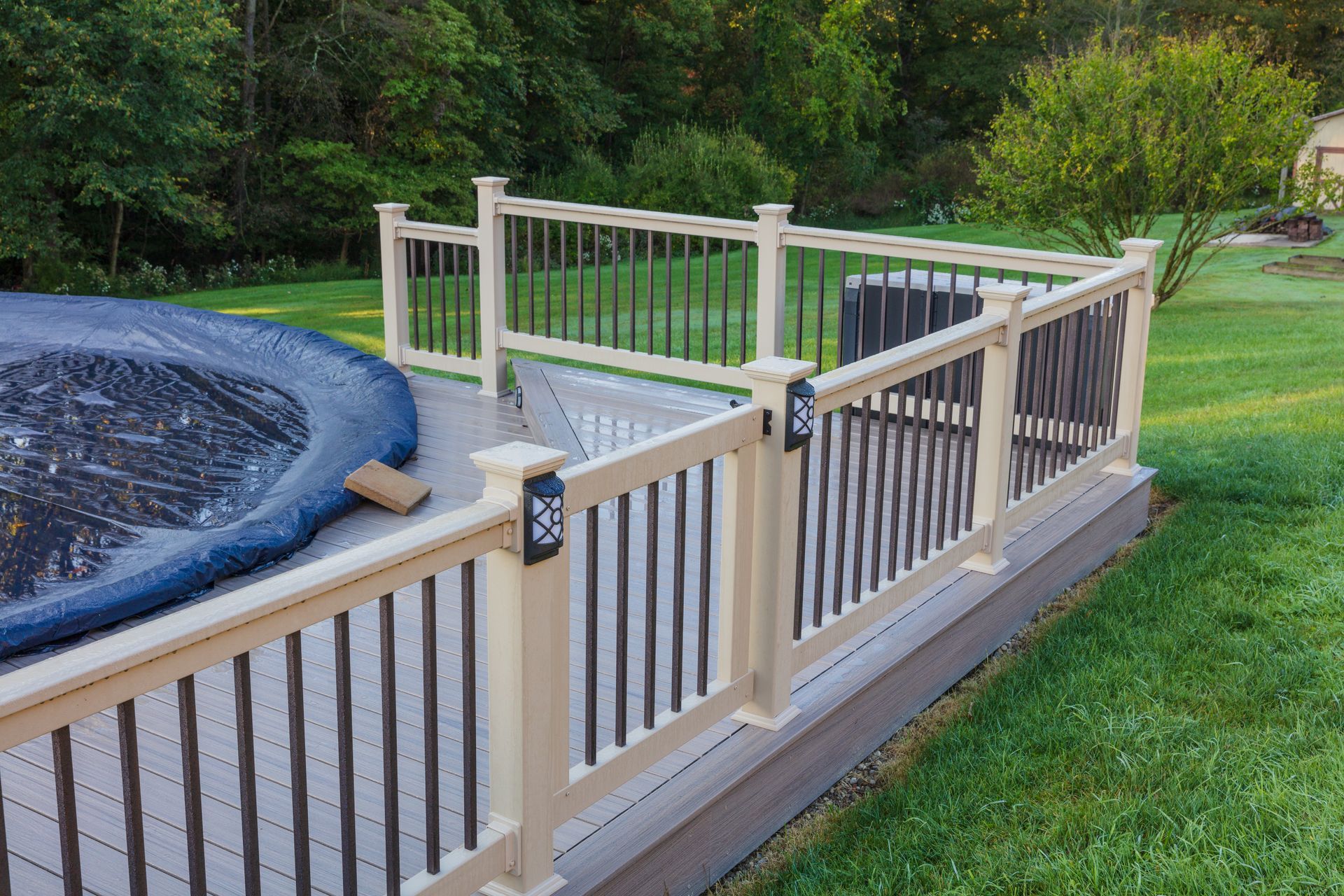 Deck with cream-colored posts and black railings, surrounding a pool covered by a blue tarp, set in a yard.
