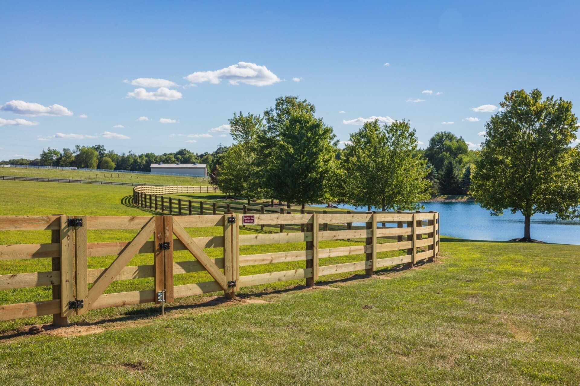 Wooden fence curves along a grassy field, with trees, a pond, and a blue sky with clouds in the background.
