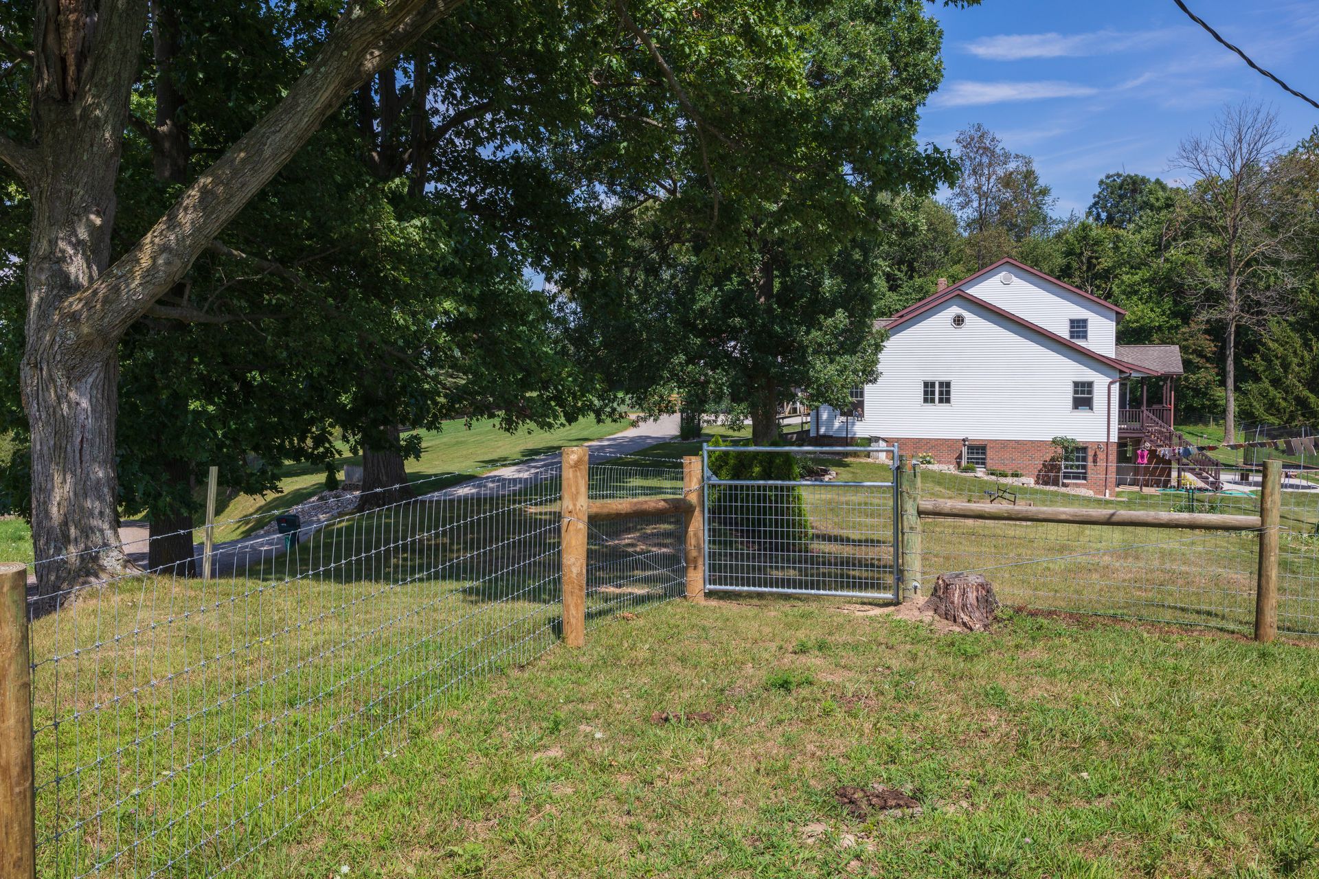 House with a white exterior sits behind a fence and green grass. Trees surround the property on a sunny day.