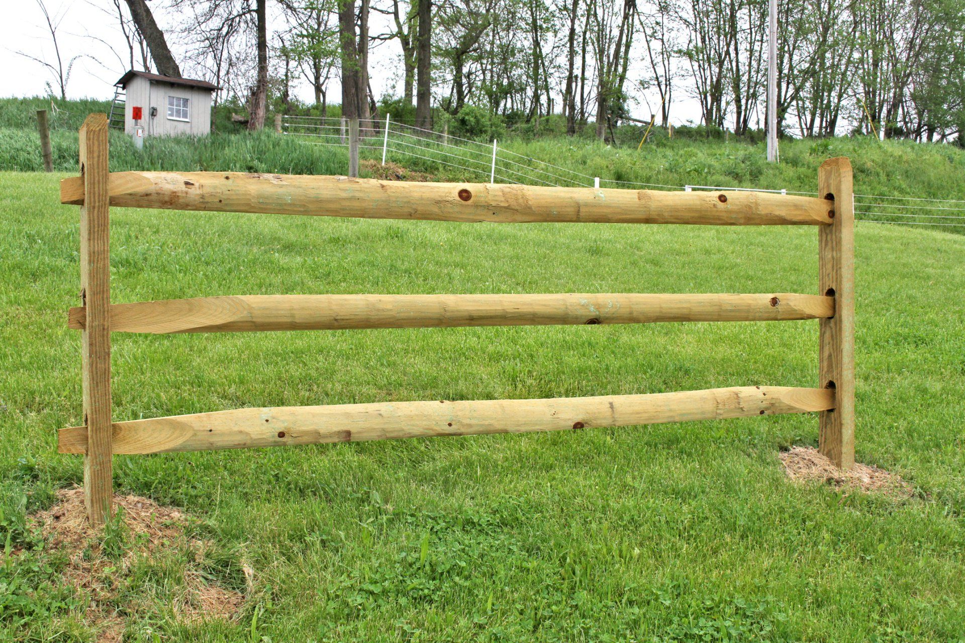 Wooden three-rail fence in a grassy field, with a small shed visible in the background.