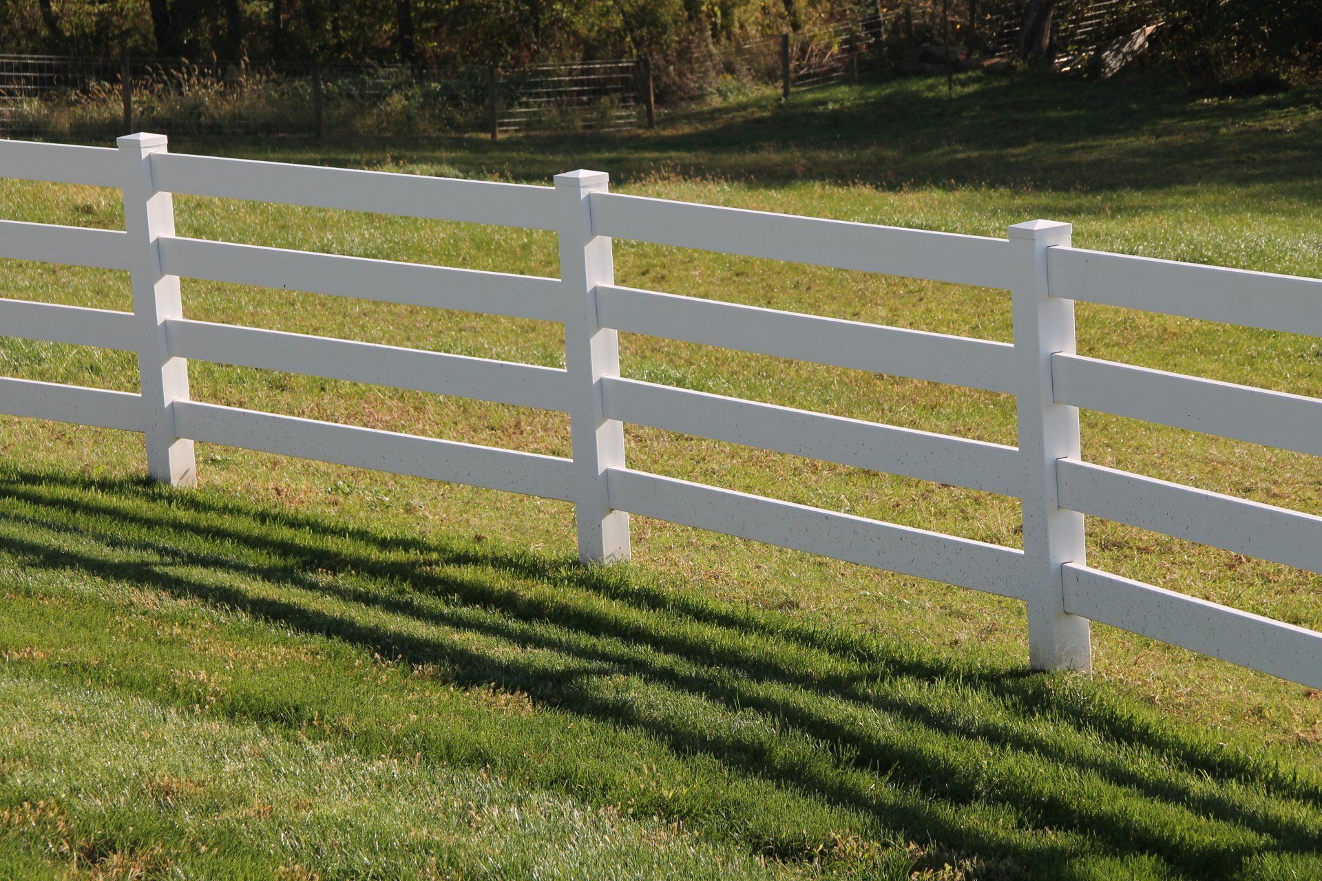 White three-rail fence in a grassy field, casting shadows on the green.