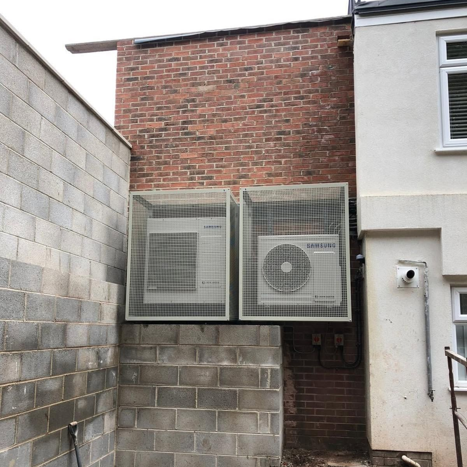 Two outdoor HVAC units mounted on a brick wall between a cinderblock wall and a house.