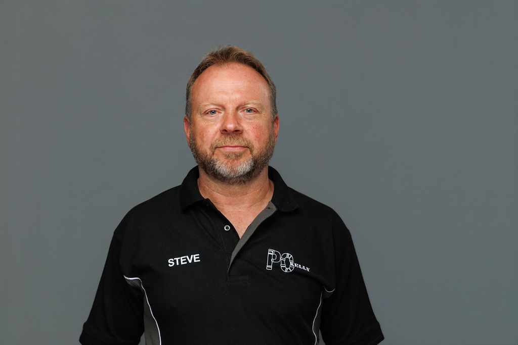 Man in a black polo shirt with logos, standing against a plain gray background.