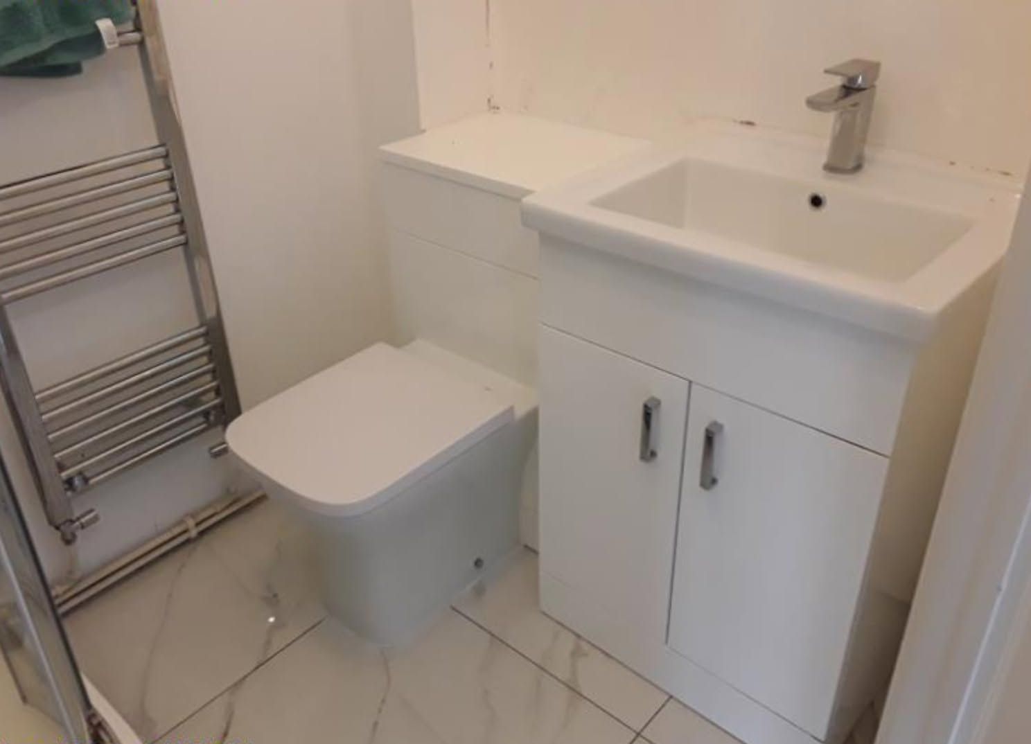 Small bathroom with white vanity, sink, toilet, and chrome towel radiator on tiled floor.
