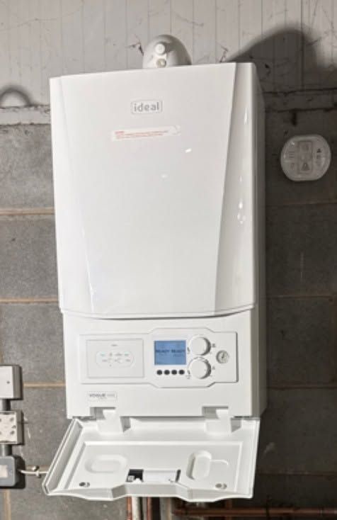 White wall-mounted boiler/control unit with open lower compartment and blue display on a gray wall.