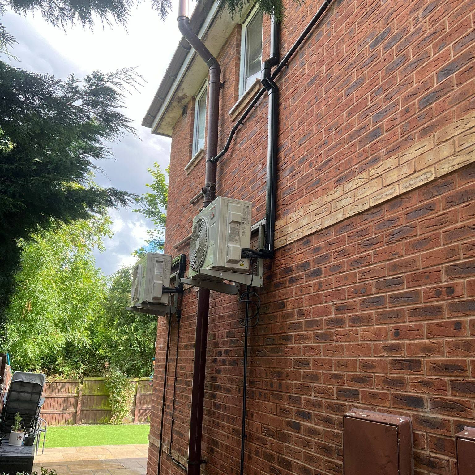 Brick apartment wall with two air-conditioning units and black pipes beside a grassy yard.