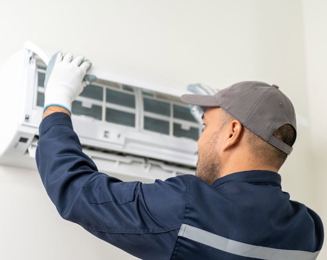 Technician cleaning a wall-mounted air conditioner with a cloth, wearing gloves and a cap.