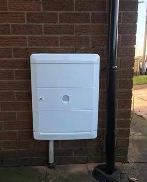 White utility box mounted on a brick wall beside a black downpipe and driveway.