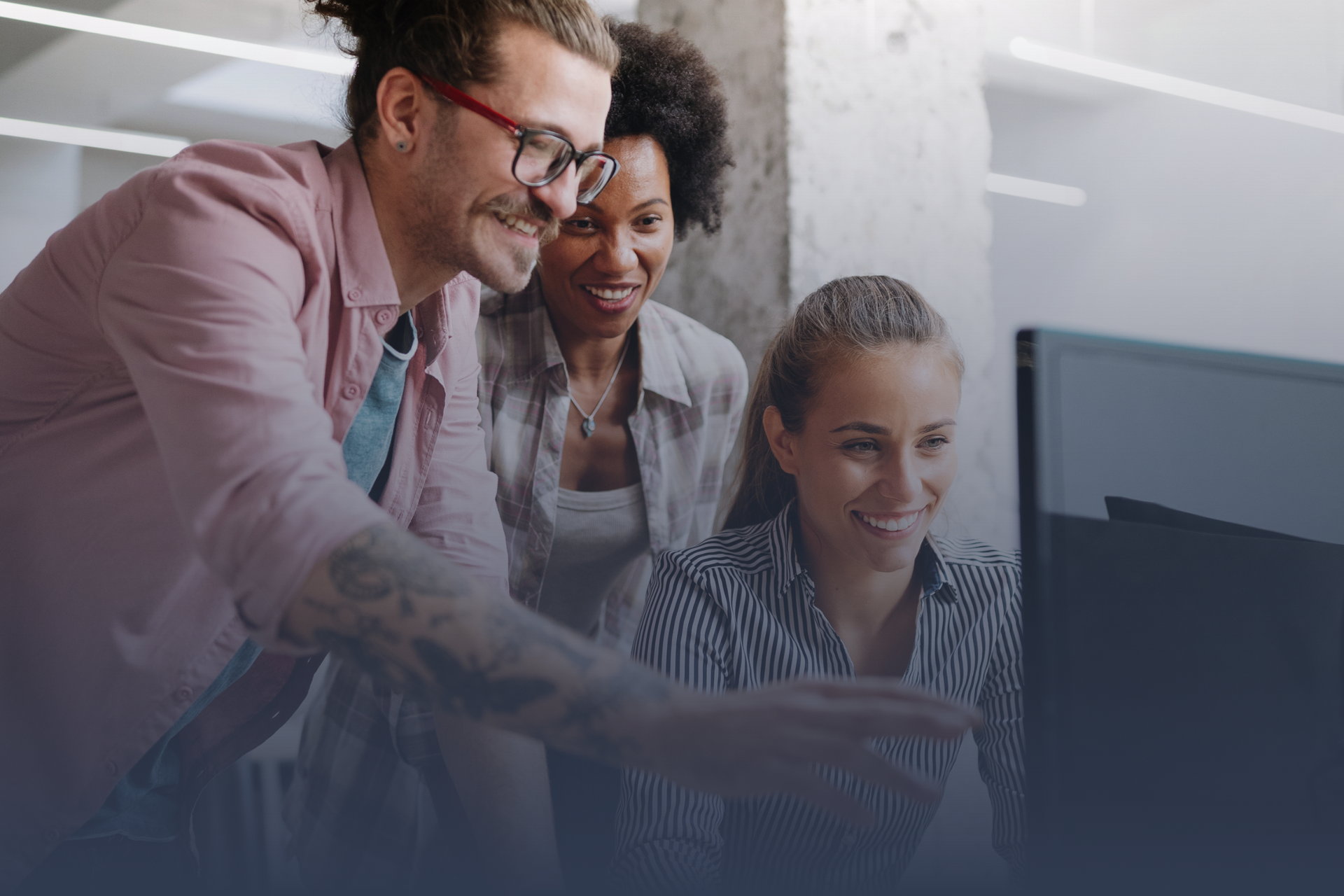 Three people looking at a computer screen, smiling. Man points with tattoo arm. Office setting.