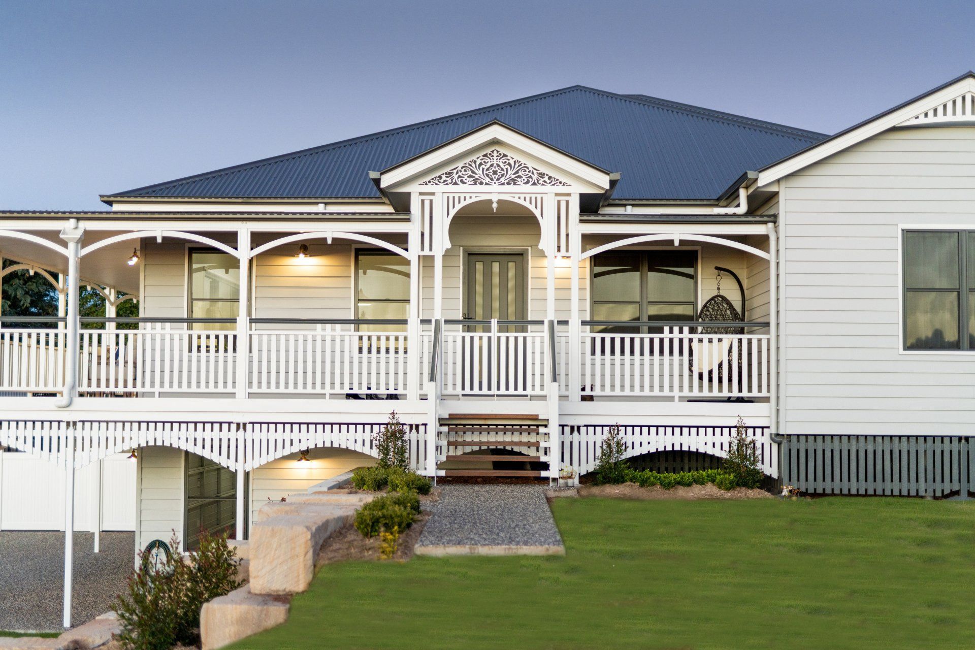 White house with a wrap-around porch and dark blue roof, set on a green lawn with a stone pathway.