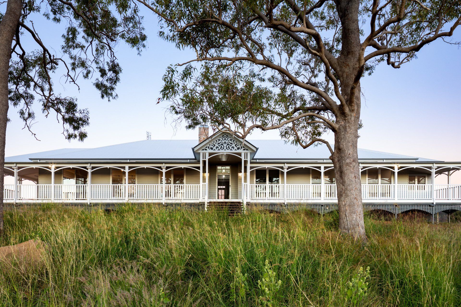 Long, light-colored house with a veranda, framed by trees and tall grass.