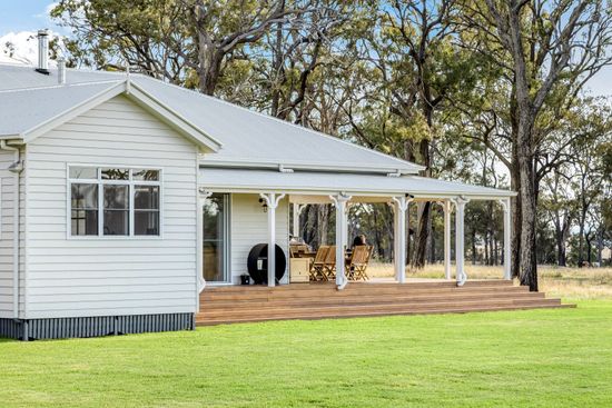 White cottage with a wraparound porch on a green lawn, surrounded by trees.