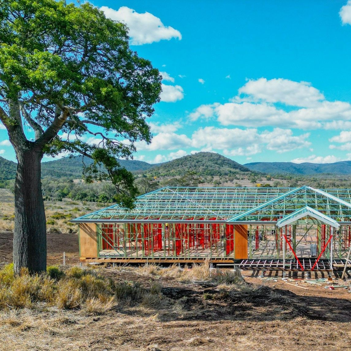 Construction site with exposed house framing against a mountain backdrop under a blue sky. A large tree stands to the left.