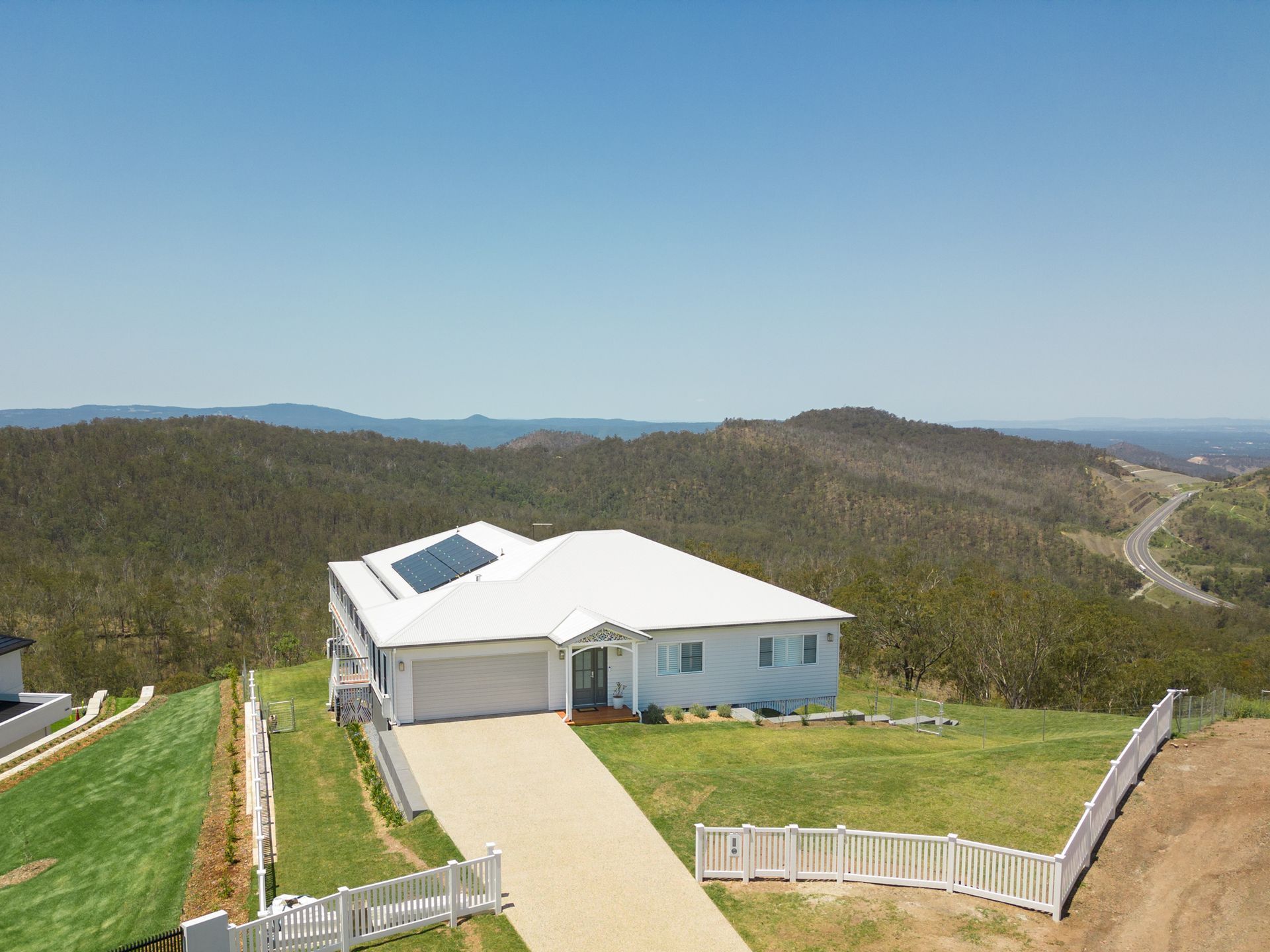 White house with solar panels on a hilltop with a driveway and white picket fence, surrounded by green hills.