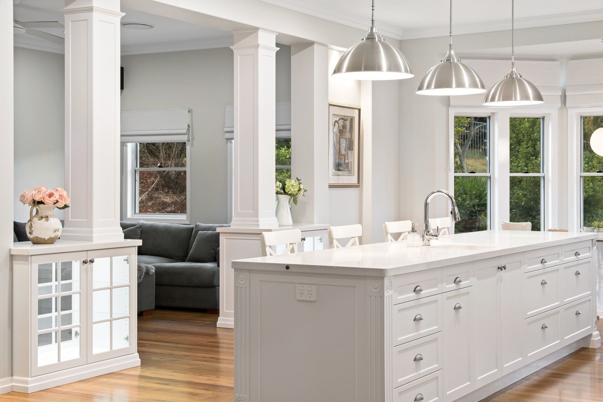Spacious white kitchen with island, pendant lights, cabinetry, and windows overlooking greenery.