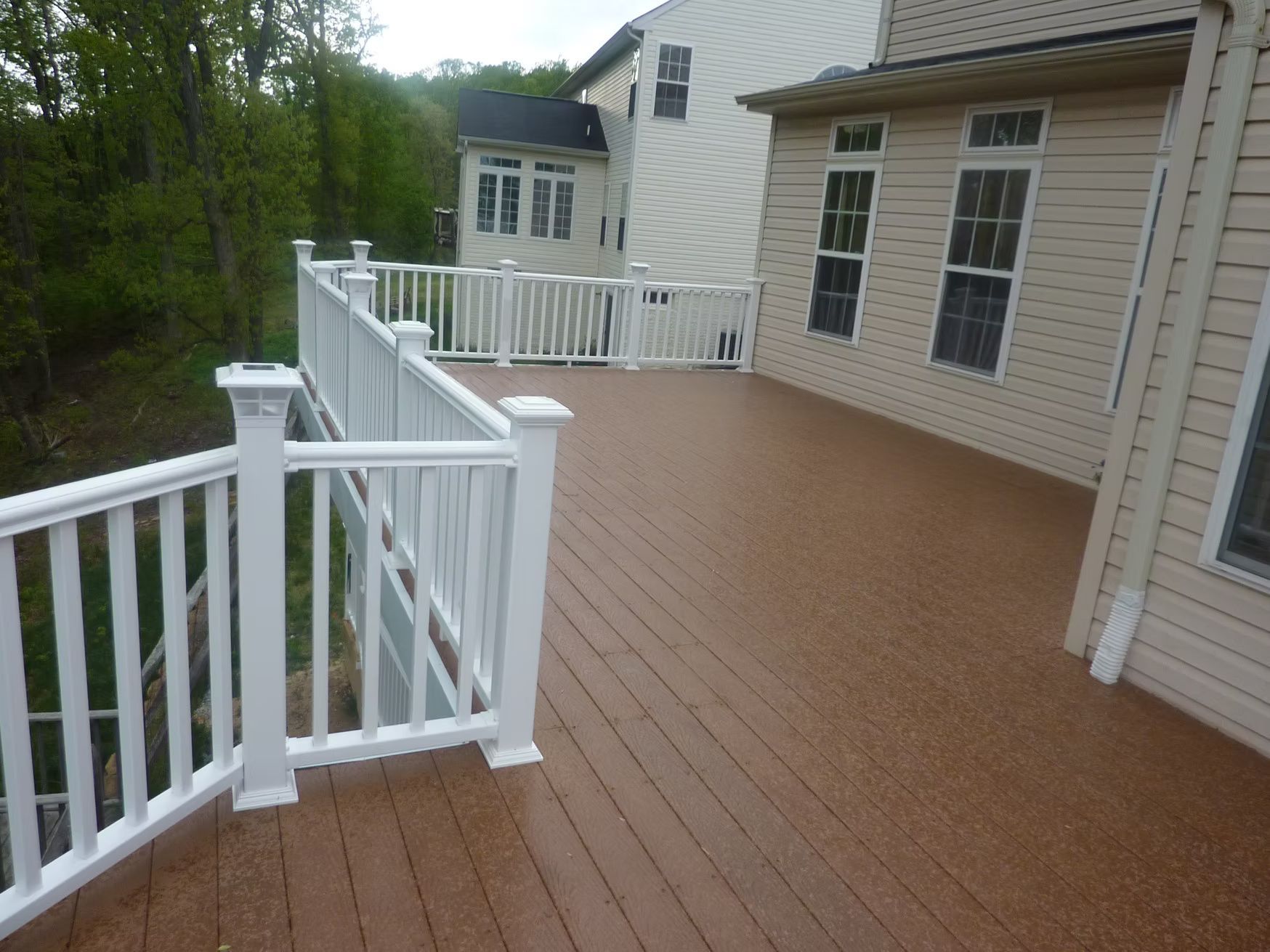 Spacious wooden deck with white railing beside a beige house and trees in the background