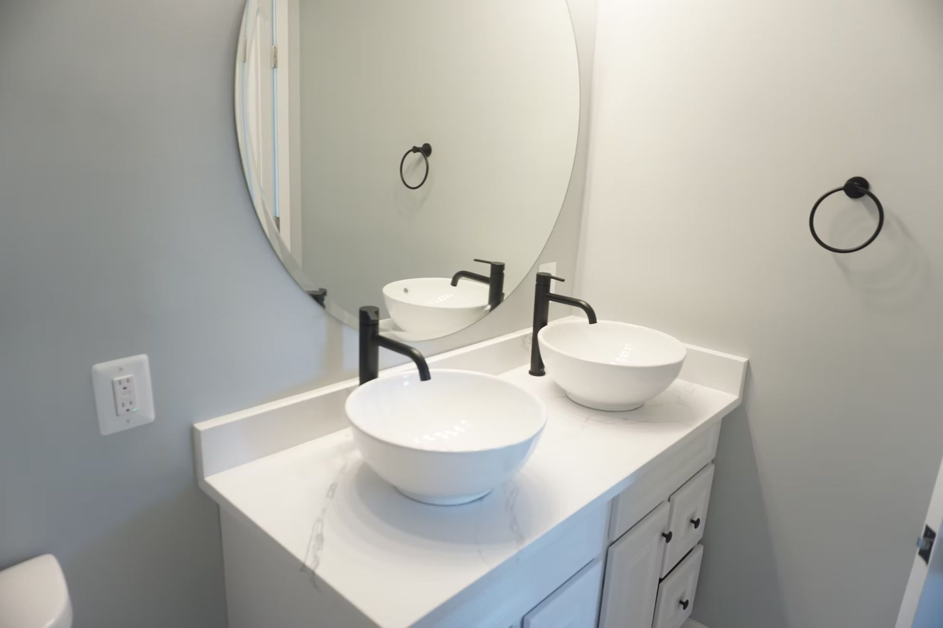 Modern bathroom vanity with white countertop, vessel sink, round mirror, and towel ring on gray walls