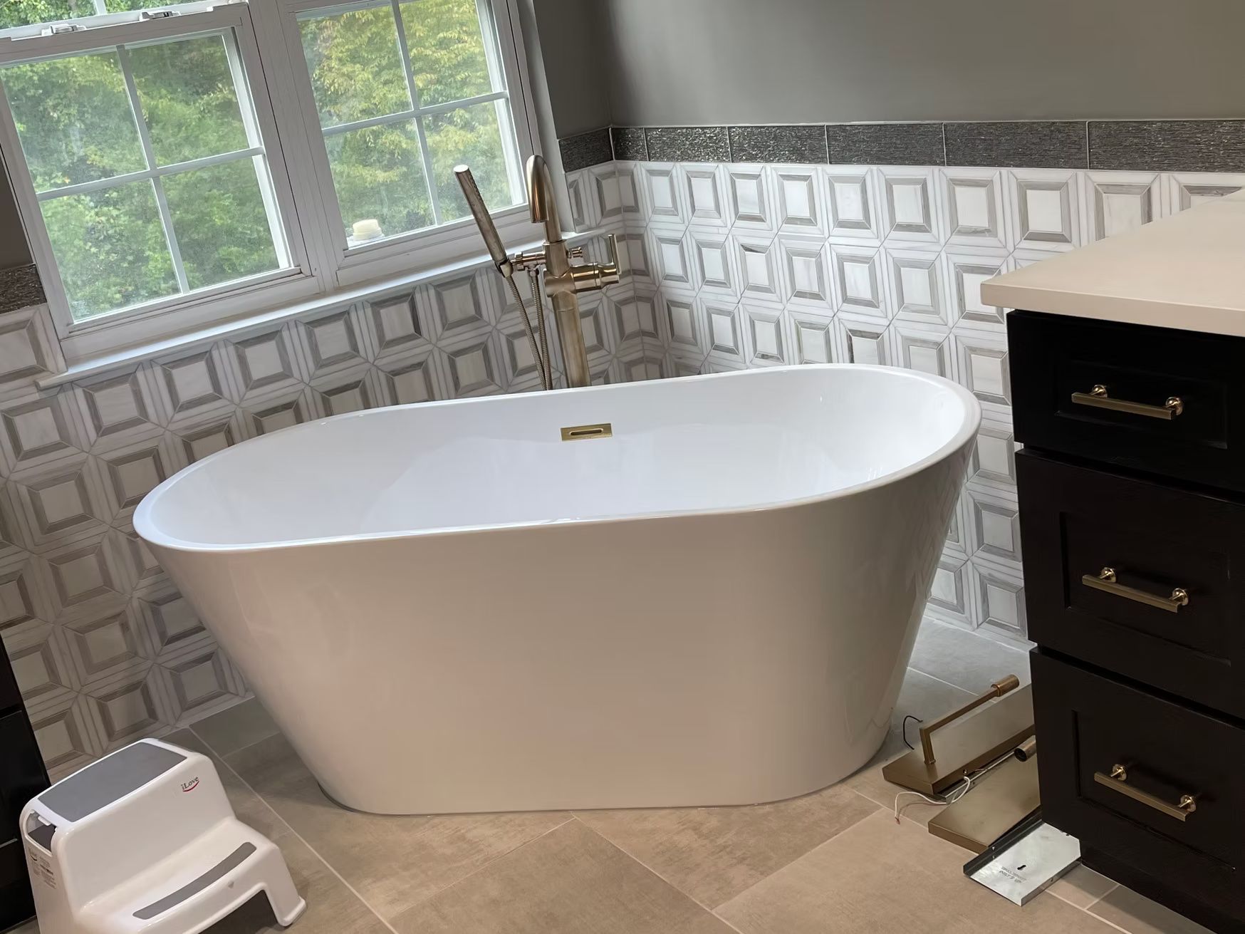Freestanding white bathtub in a tiled bathroom beside a window and dark vanity cabinet