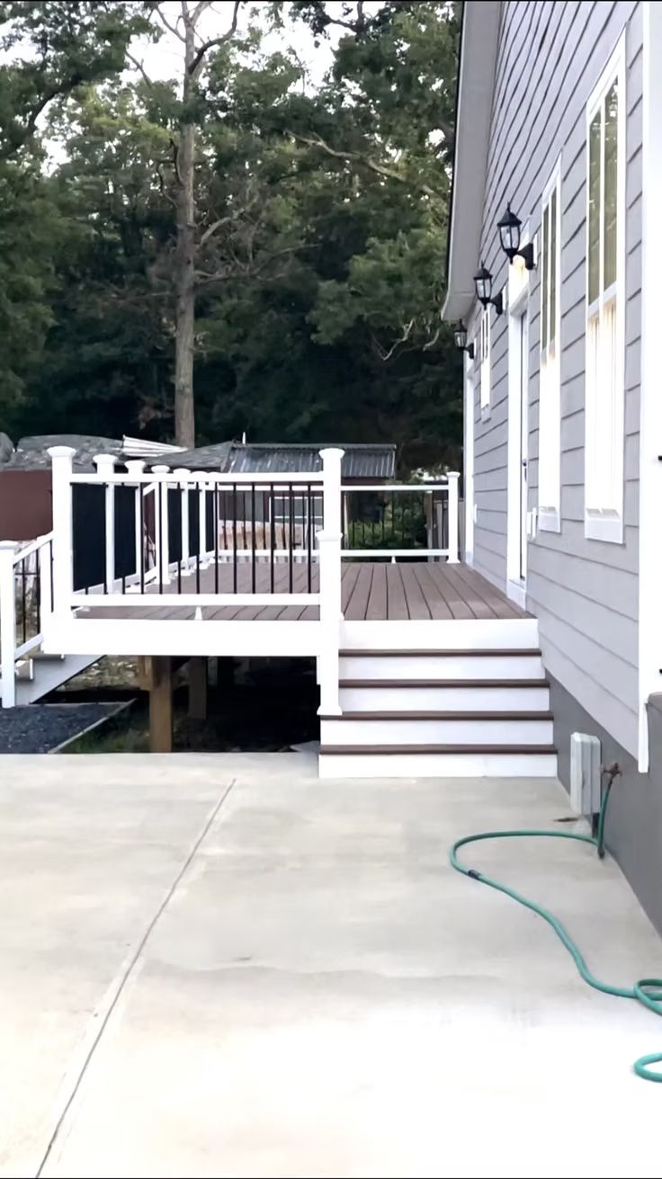 White deck stairs beside a gray house with a hose on a concrete patio and trees in the background