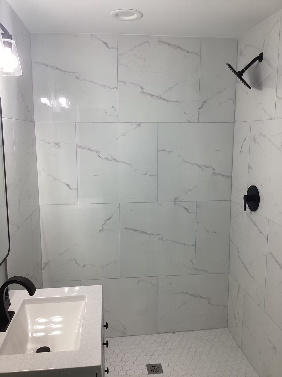 Modern tiled bathroom shower with marble-look walls, sink, black fixtures, and a handheld showerhead.
