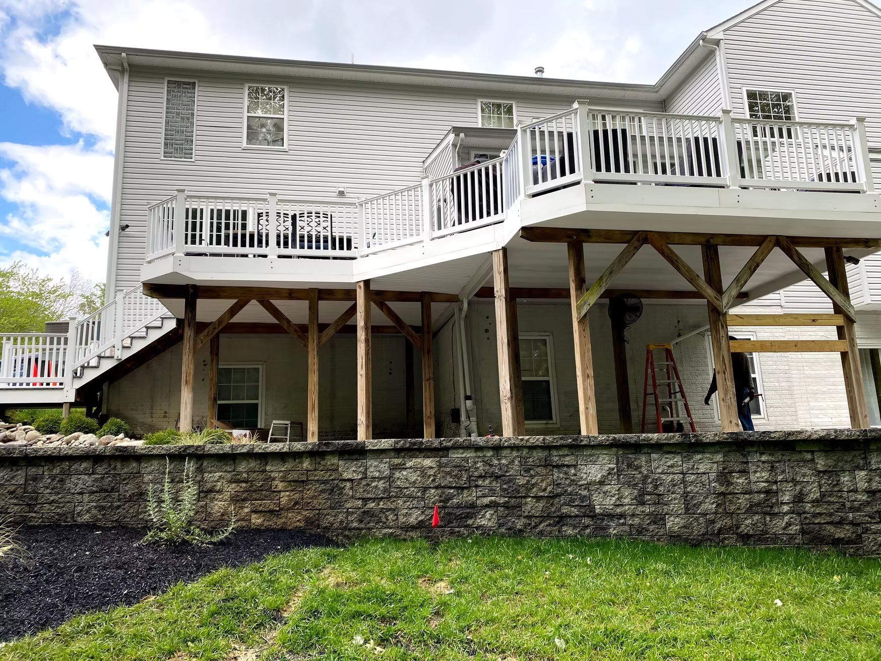 White two-story house with raised deck, stone retaining wall, and green lawn underneath