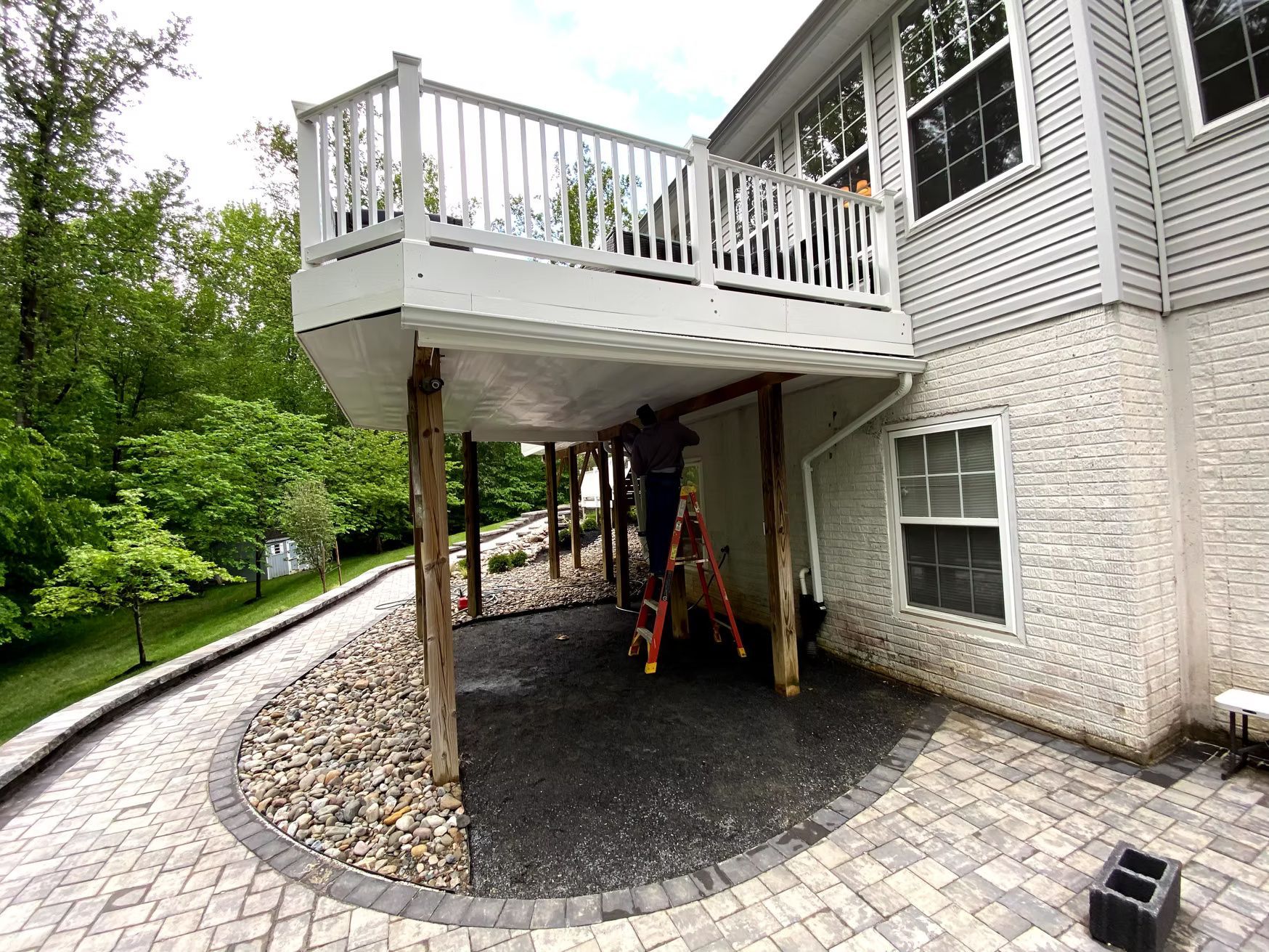 Raised deck over stone patio with support posts beside a house and garden path
