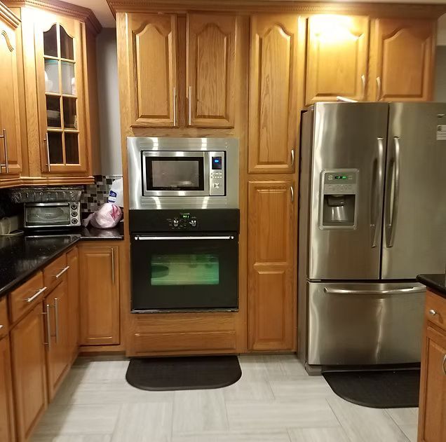 Wooden kitchen with stainless steel refrigerator, built-in oven and microwave, and black countertops.