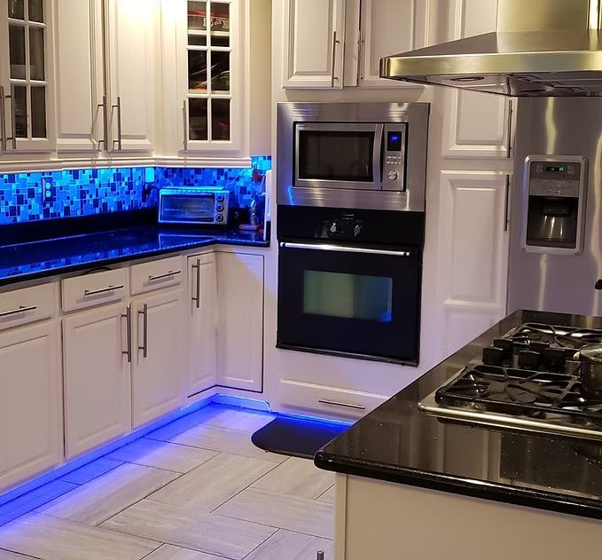 Modern kitchen with white cabinets, blue under-cabinet lighting, black countertops, and stainless steel appliances
