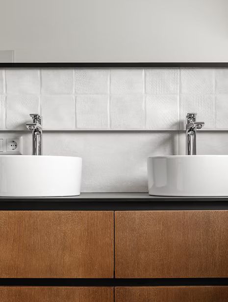 Modern bathroom sink with white basins, chrome faucets, tiled wall, and wood vanity cabinet