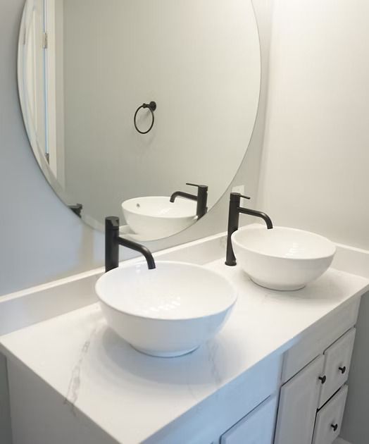 Modern bathroom vanity with two white vessel sinks and black faucets beneath a round mirror