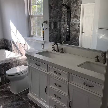 Modern bathroom with gray vanity, large mirror, double sinks, and a toilet by the window.