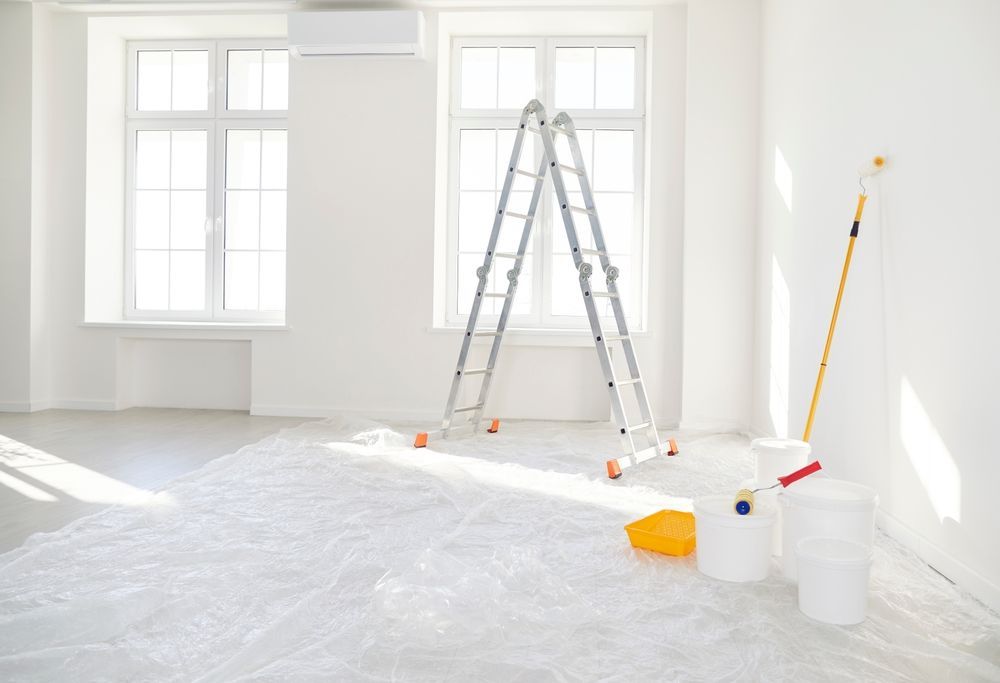 Bright room with plastic-covered floor, ladder, paint supplies, and windows during renovation.