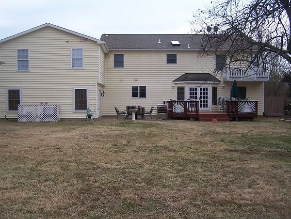Backyard view of a beige two-story house with a wooden deck and patio furniture under bare trees