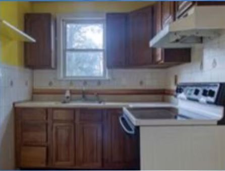 Small kitchen with wooden cabinets, white stove, sink under a window, and tiled walls.