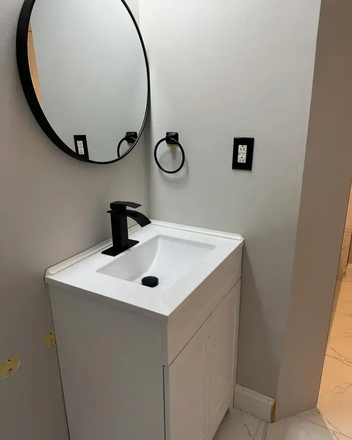 Modern bathroom vanity with white sink, black faucet, round mirror, and wall outlet.