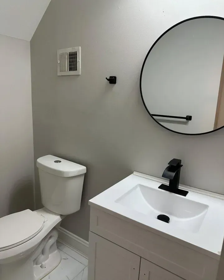 Small white bathroom with toilet, vanity sink, round mirror, and gray walls