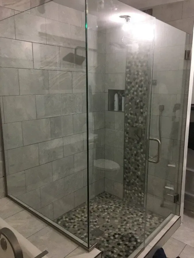 Glass walk-in shower with gray tile walls, pebble floor, and a chrome handle