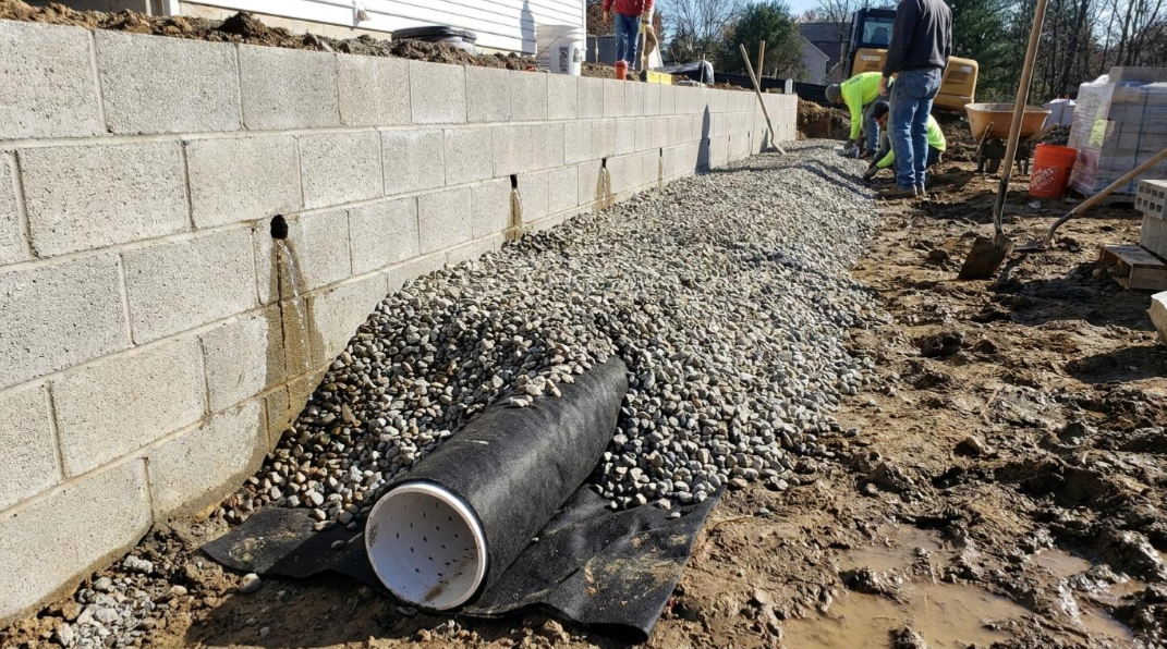 Construction site with a black pipe in gravel beside a cinder block wall and workers in the background