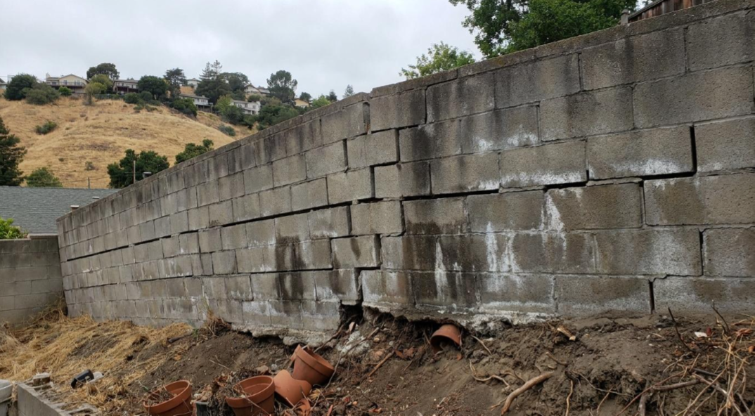 Cracked concrete retaining wall on a muddy slope with debris and broken pipes at the base
