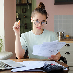 Woman with glasses studying documents, calculator, and laptop on a table in a kitchen.