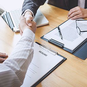 Two people shaking hands over a wooden table, with a laptop, resume, and documents visible.