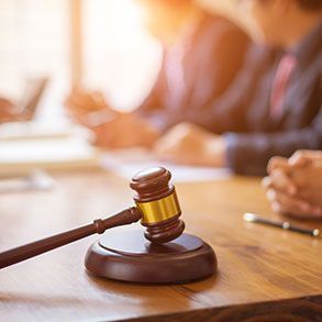 Gavel on wooden table with people blurred in the background, lit by sunlight.