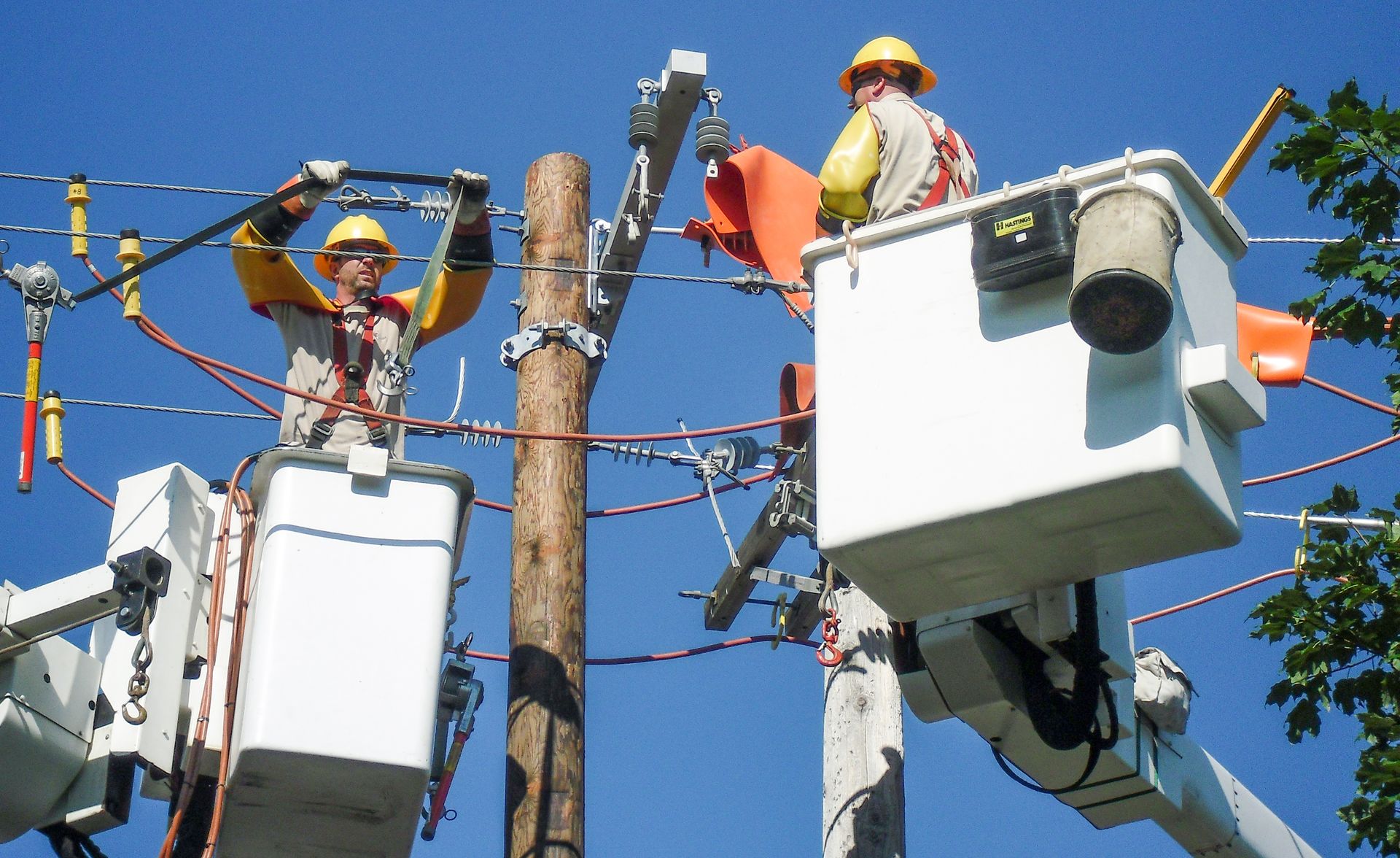 Two utility workers in bucket trucks repairing power lines on a wooden pole. Bright blue sky.