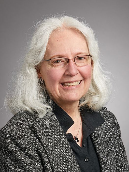 Woman with gray hair and glasses smiles, wearing a black top and gray patterned jacket.