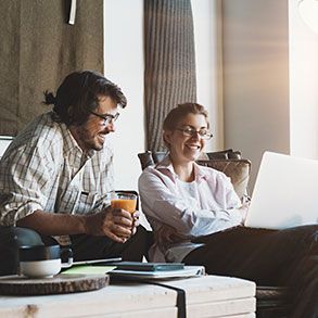 Two people looking at laptop, smiling. Man holds drink. Seated in a living room.