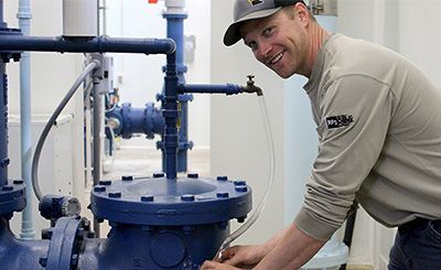 Man in a cap smiles while working on blue pipes with a water tap in a utility room.