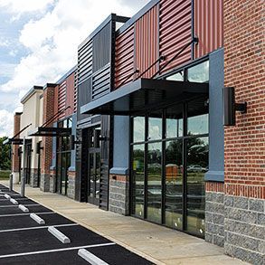 Strip mall storefronts with red corrugated metal, brick, and gray accents. Dark awnings and parking spaces.