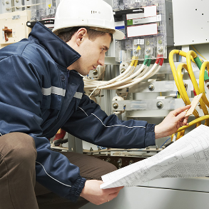 Electrician in hard hat examines wiring diagram, working on an electrical panel.