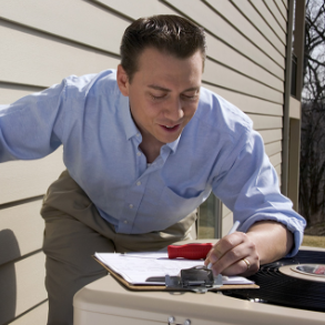 Man in blue shirt inspecting air conditioner, taking notes outdoors.