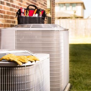 Two air conditioning units outdoors with a toolbox and work gloves, near a brick wall and grass.